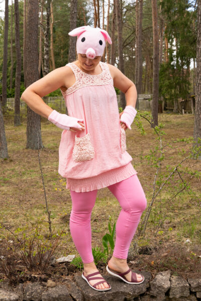 Roosa notsu kostüüm naise seljas. Kostüümi kuulub kleit, kindad, peakate ja jalas on roosad retuusid. Naine seisab kivimüüri peal, taga on männid.