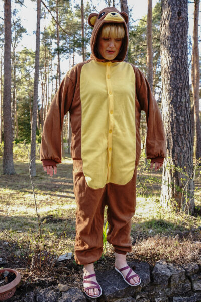 Karu kostüüm naise seiljas. Karu on pruun, kollase kõhuga, kombe on kapuutsiga. Karu seisab männipuude ees.