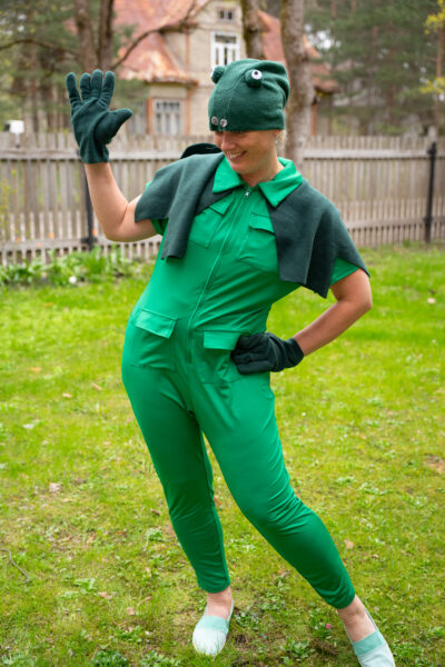 Naine rohelises kombinesoonis, peas roheline konna silmadega müts aj käes rohelised sõrmkidad. NAine seisab õues, muru on roheline ja taga on puust aed.