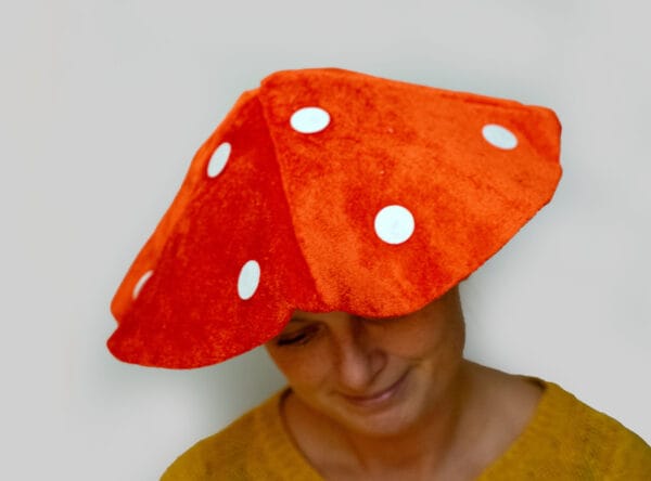 A person wearing a playful red and white spotted mushroom hat (toadstool style) and a mustard yellow sweater, photographed against a blurred forest background with natural lighting filtering through the trees, creating a whimsical woodland scene.