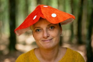 A person wearing a playful red and white spotted mushroom hat (toadstool style) and a mustard yellow sweater, photographed against a blurred forest background with natural lighting filtering through the trees, creating a whimsical woodland scene. Inimene kannab kärbseseene mütsi.