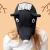Handmade black bear costume hat made of fleece, featuring a unique white fringe on top and a white nose. A model is wearing the hat against a peach-colored background with hand-drawn branch illustrations.