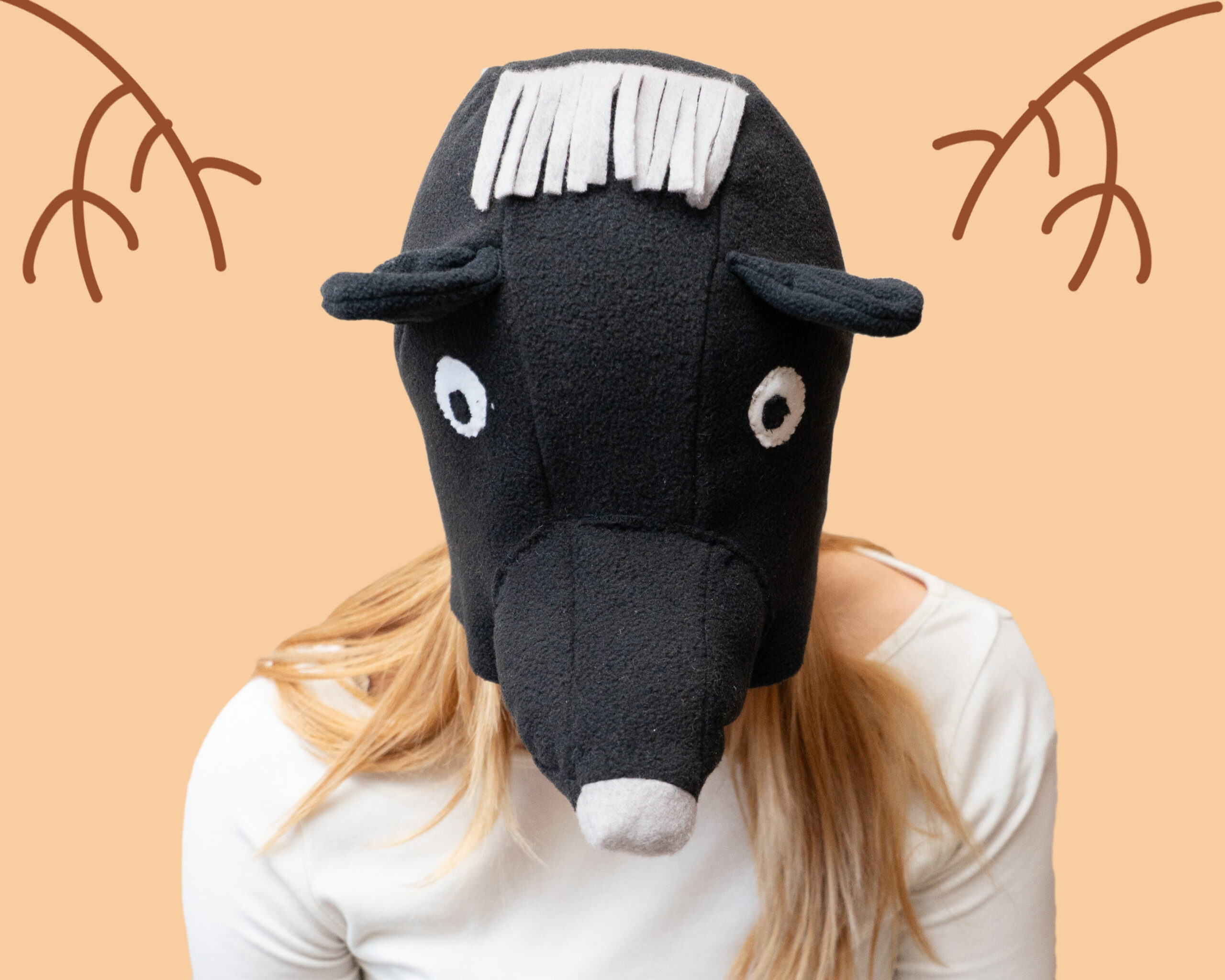 Handmade black bear costume hat made of fleece, featuring a unique white fringe on top and a white nose. A model is wearing the hat against a peach-colored background with hand-drawn branch illustrations.