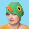 Handmade vibrant green fleece bird hat for adults with an orange beak and large yellow-black eyes. A woman is modeling the hat against a bright blue background.