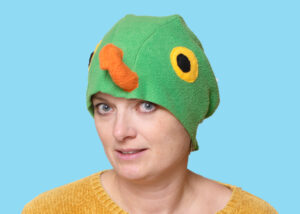 Handmade vibrant green fleece bird hat for adults with an orange beak and large yellow-black eyes. A woman is modeling the hat against a bright blue background.