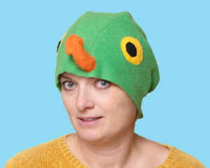 Handmade vibrant green fleece bird hat for adults with an orange beak and large yellow-black eyes. A woman is modeling the hat against a bright blue background.