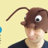 Handmade brown fleece wood ant costume hat for adults. The hat features large white eyes, brown mandibles, and long wired antennae. A man is wearing the hat against a light yellow background next to a stylized blue flower illustration.