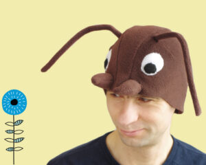 Handmade brown fleece wood ant costume hat for adults. The hat features large white eyes, brown mandibles, and long wired antennae. A man is wearing the hat against a light yellow background next to a stylized blue flower illustration.