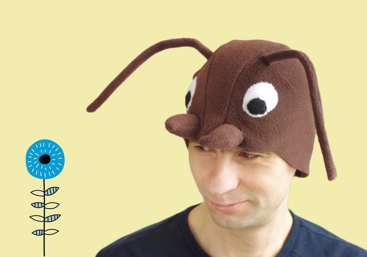 Handmade brown fleece wood ant costume hat for adults. The hat features large white eyes, brown mandibles, and long wired antennae. A man is wearing the hat against a light yellow background next to a stylized blue flower illustration.