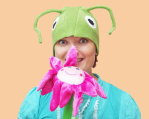 Naine kannab suurte valgete silmade ja pikkade tundlatega helerohelise rohutirtsu kostüümi- Käes on suur neoonroosa naerunäoga lill.