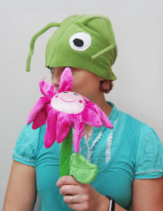 Naine kannab suurte valgete silmade ja pikkade tundlatega helerohelise rohutirtsu kostüümi- Käes on suur neoonroosa naerunäoga lill.